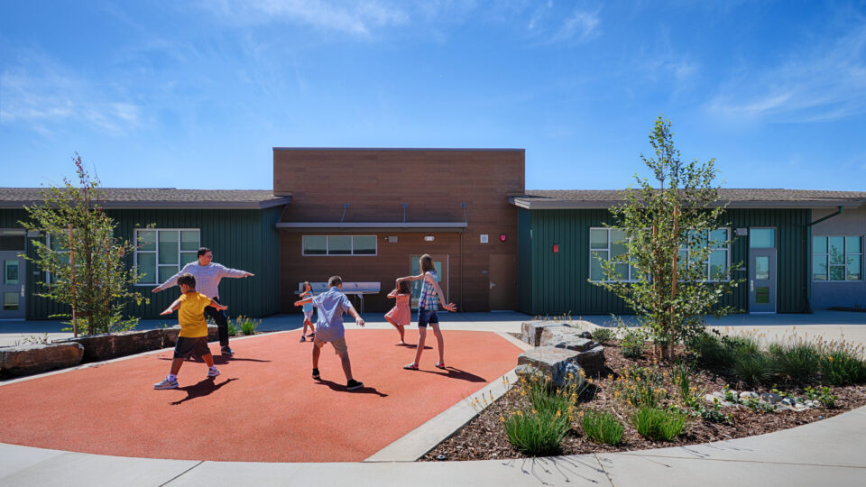 Children and an adult play on a red courtyard at a modern school building under a bright blue sky