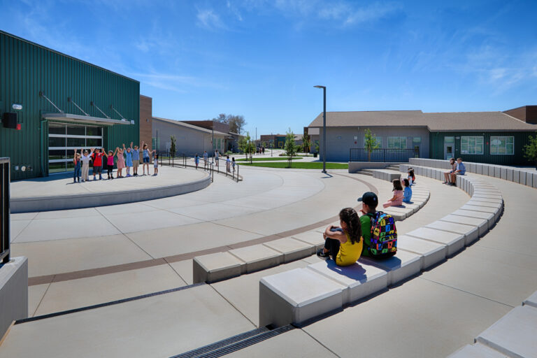 Children gathered in a school courtyard with curved benches, some sitting and others waving near modern buildings under a blue sky