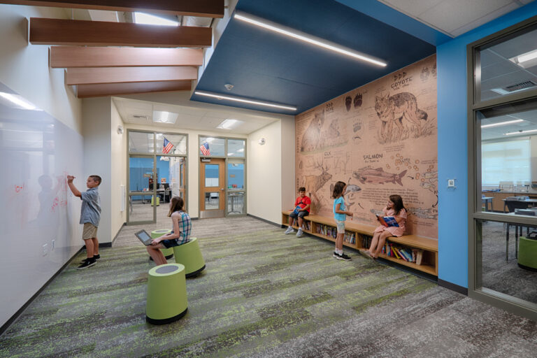 Children use a modern school hallway, with one writing on a glass wall, one using a tablet, and others reading by a wildlife mural