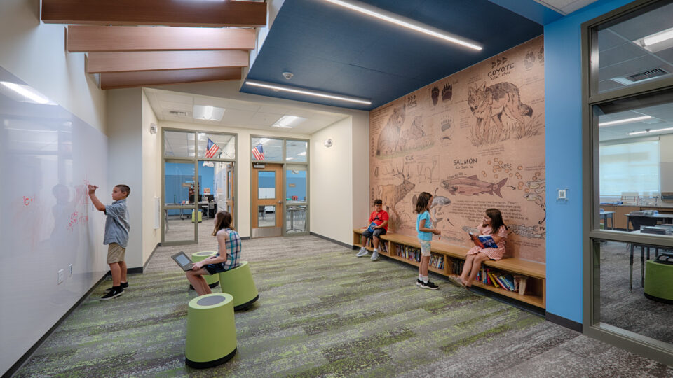 Children use a modern school hallway, with one writing on a glass wall, one using a tablet, and others reading by a wildlife mural