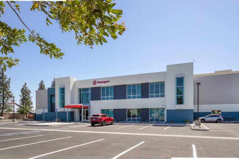 Modern industrial building with a Theragent sign and mostly empty parking lot under a clear blue sky
