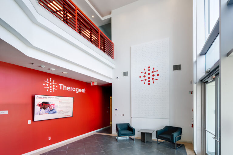 Modern lobby with a red Theragent wall, two teal chairs, and a tall white atrium with red railing accents