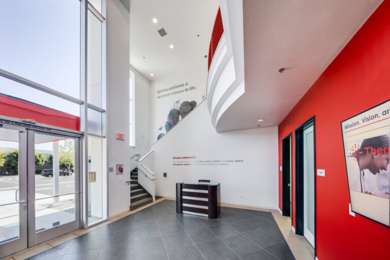 Bright modern office lobby with tall glass windows, red accent walls, staircase, and a small reception desk