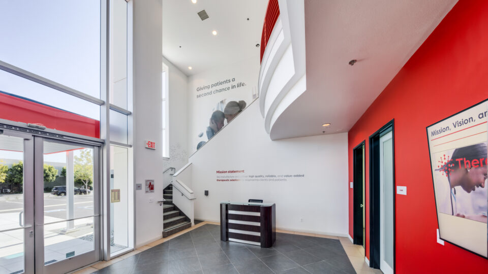 Bright modern office lobby with tall glass windows, red accent walls, staircase, and a small reception desk