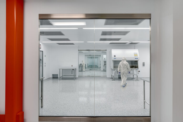 Cleanroom laboratory seen through glass doors, with workers in protective suits and a bright red column in the foreground
