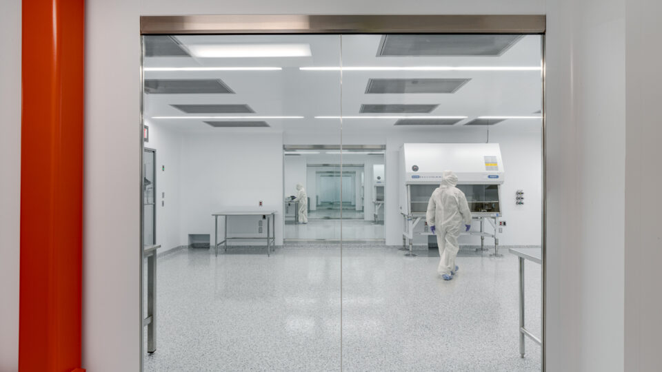 Cleanroom laboratory seen through glass doors, with workers in protective suits and a bright red column in the foreground