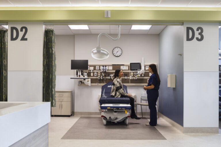 Patient sitting on an exam bed in a medical bay while a nurse talks with her