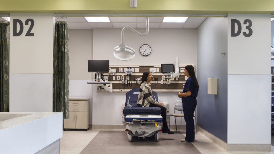 Patient sitting on an exam bed in a medical bay while a nurse talks with her