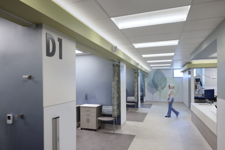 Modern hospital hallway with private exam rooms labeled D1 and D2, a nurse walking by, and medical equipment nearby