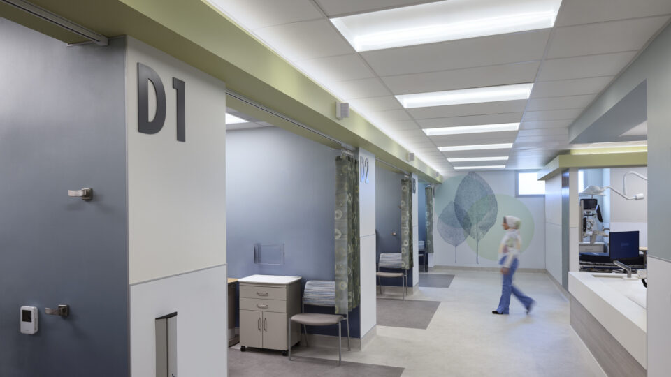 Modern hospital hallway with private exam rooms labeled D1 and D2, a nurse walking by, and medical equipment nearby