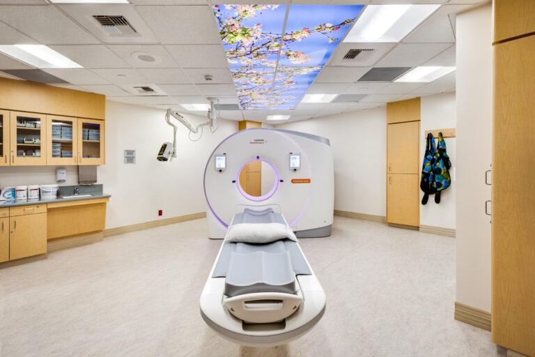 Hospital imaging room with a CT scanner and patient table under a ceiling panel showing cherry blossoms