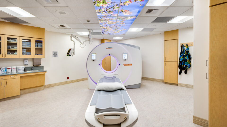 Hospital imaging room with a CT scanner and patient table under a ceiling panel showing cherry blossoms