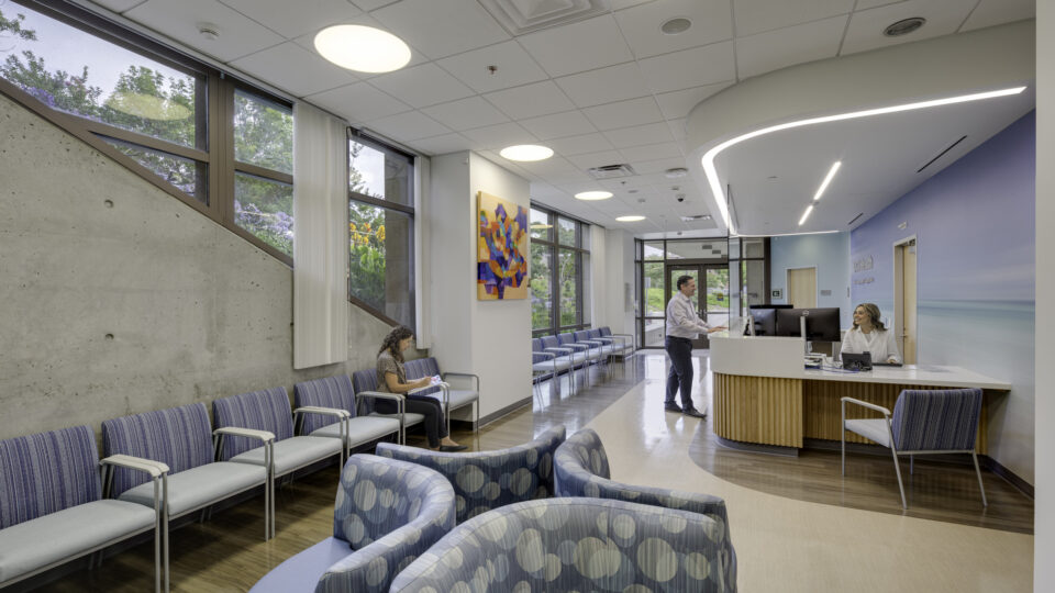 Modern medical waiting room with reception desk, blue seating, large windows, and three people inside