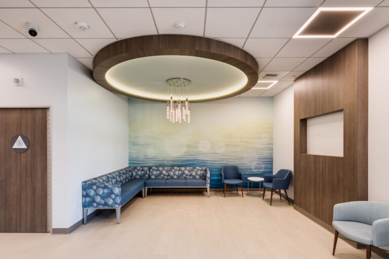 Modern waiting area with blue sectional seating, two chairs, a small table, and a circular chandelier under a wood ceiling feature