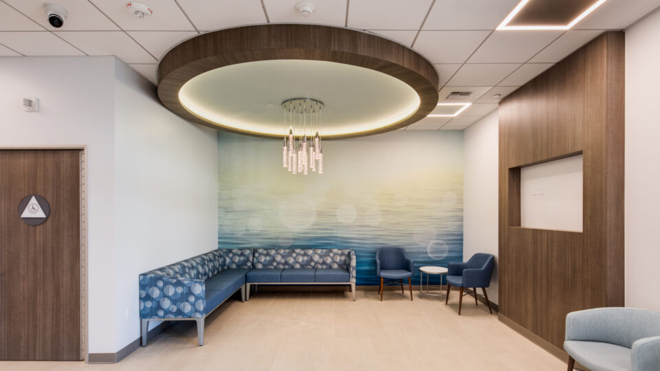 Modern waiting area with blue sectional seating, two chairs, a small table, and a circular chandelier under a wood ceiling feature