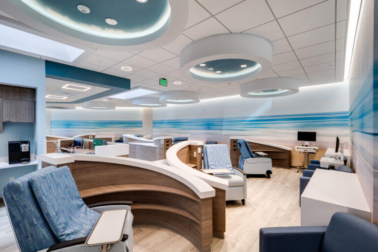 Modern hospital infusion room with curved wooden cubicles, blue reclining chairs, and ceiling ring lights with a beach mural wall