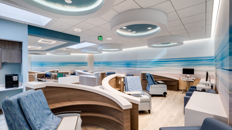 Modern hospital infusion room with curved wooden cubicles, blue reclining chairs, and ceiling ring lights with a beach mural wall
