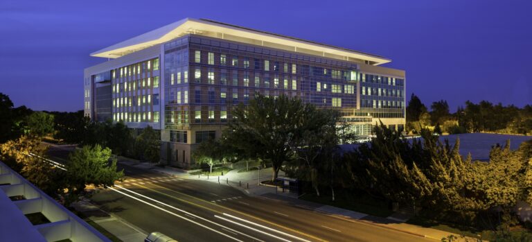 Modern glass office building at dusk with illuminated windows, trees, and light trails on the street below