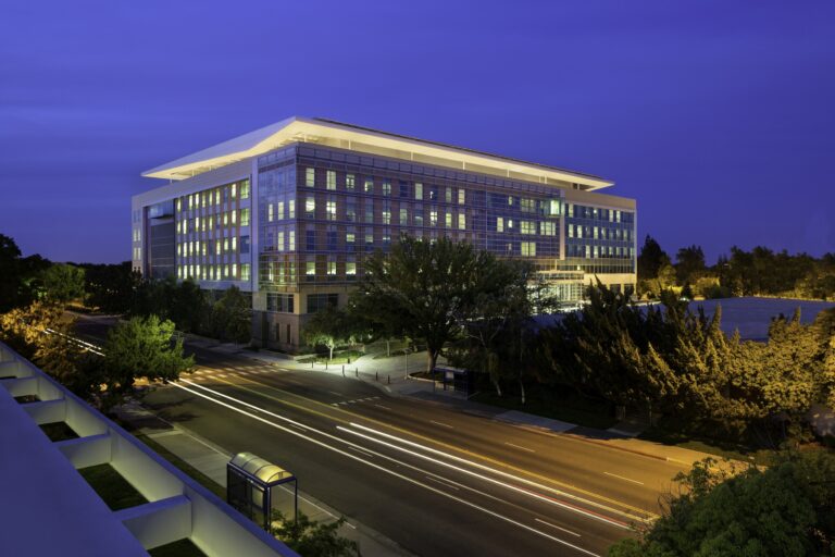 Modern glass office building at dusk with illuminated windows, trees, and light trails on the street below