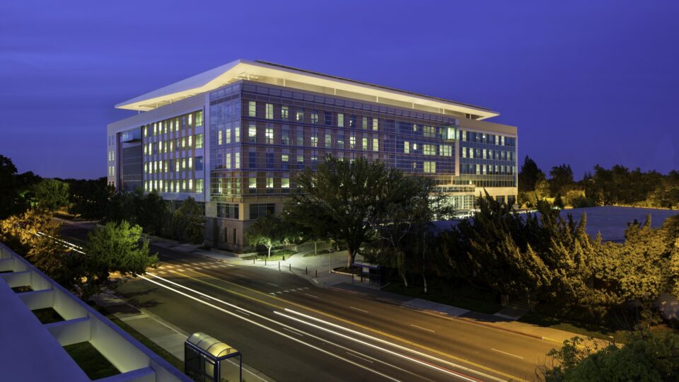 Modern glass office building at dusk with illuminated windows, trees, and light trails on the street below