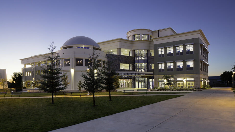 Modern science center and museum building at dusk with a dome, lit windows, and a curved walkway in front