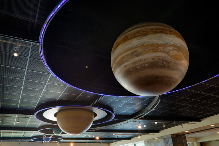 Suspended model planets, including Jupiter, hang from a dark ceiling with purple-lit rings in a science museum-like space