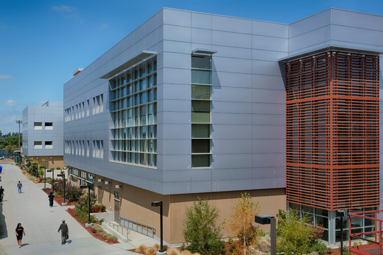 Modern campus building with gray panels and wood slats, beside a walkway with a few people under a blue sky