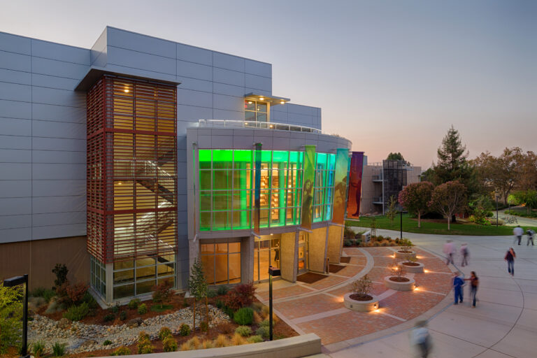 Modern campus building at dusk with colorful glass facade, lit windows, and people walking outside