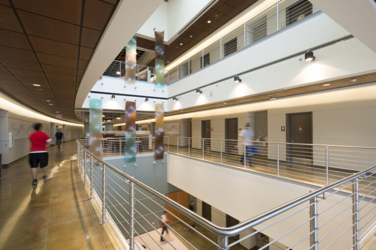 Modern indoor atrium hallway with railings, colorful hanging panels, and blurred people walking past restroom doors