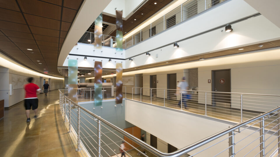 Modern indoor atrium hallway with railings, colorful hanging panels, and blurred people walking past restroom doors