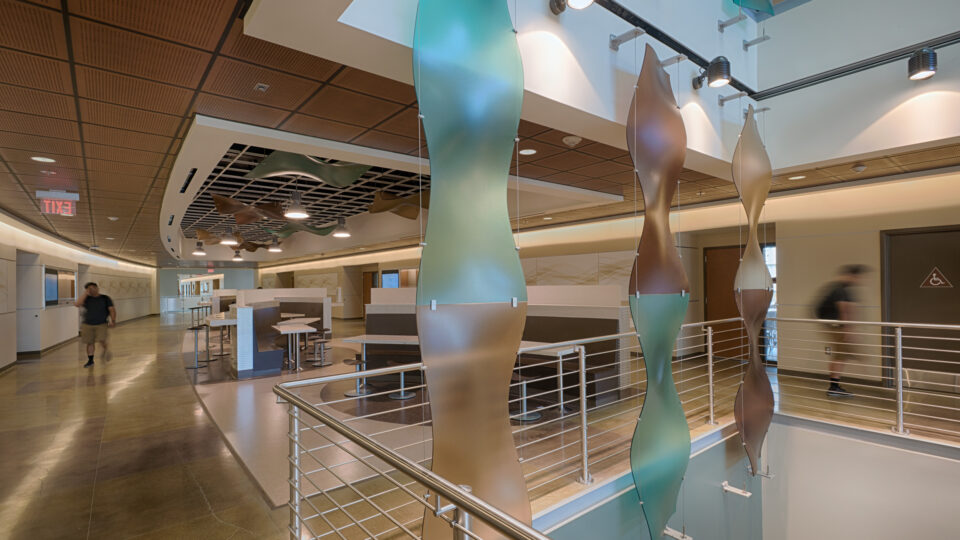 Modern hospital corridor with abstract hanging panels, railings, seating, and a few blurred people walking