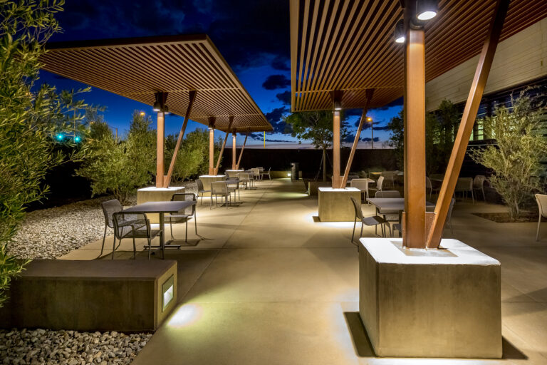 Modern outdoor patio with tables and chairs under lit wood-slat canopies at dusk