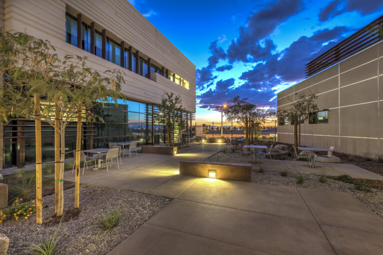 Modern outdoor courtyard between office buildings at dusk with lit walkways, tables, trees, and a dramatic blue sunset sky