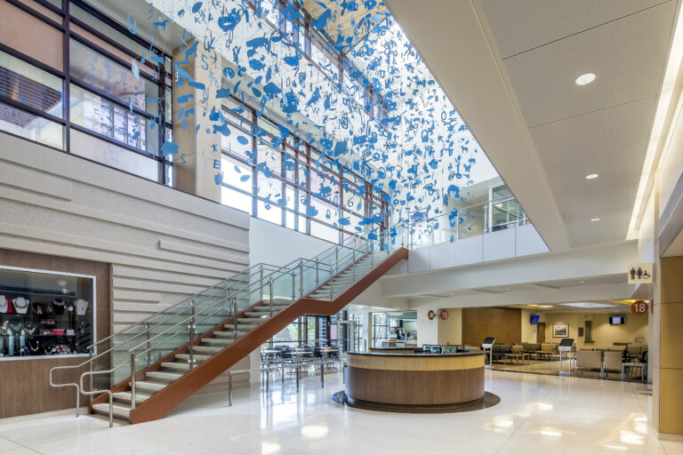Modern lobby with a staircase, information desk, and hanging blue letter-like art beneath a skylit ceiling