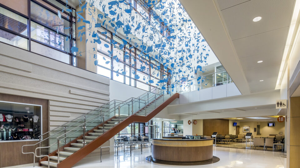 Modern lobby with a staircase, information desk, and hanging blue letter-like art beneath a skylit ceiling