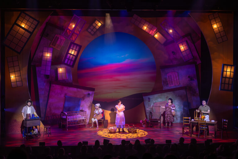 Theater stage with five actors in a colorful village set, lit by magenta and blue lights with a sunset backdrop