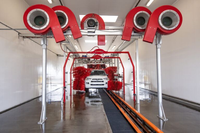 White SUV being cleaned in an automated car wash with large red brushes and overhead dryers