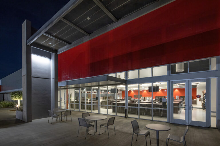Modern building exterior at night with a red facade, glass walls, and patio tables outside a brightly lit interior