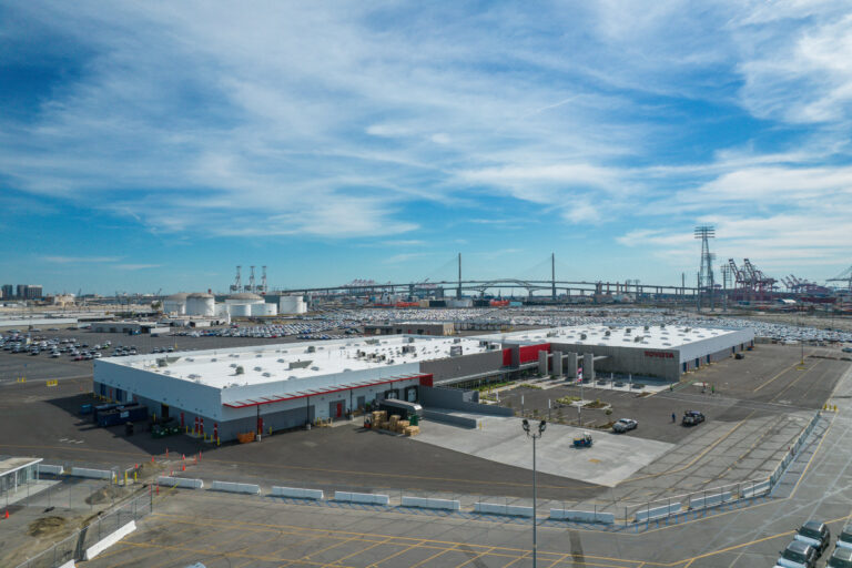 Industrial port with Toyota buildings, parking lots, cranes, and a cable-stayed bridge under a cloudy blue sky
