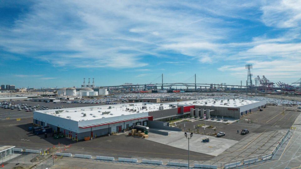 Industrial port with Toyota buildings, parking lots, cranes, and a cable-stayed bridge under a cloudy blue sky