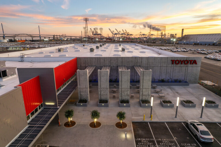 Toyota facility with rooftop, parking lot, and shipping cranes at sunset in an industrial port setting