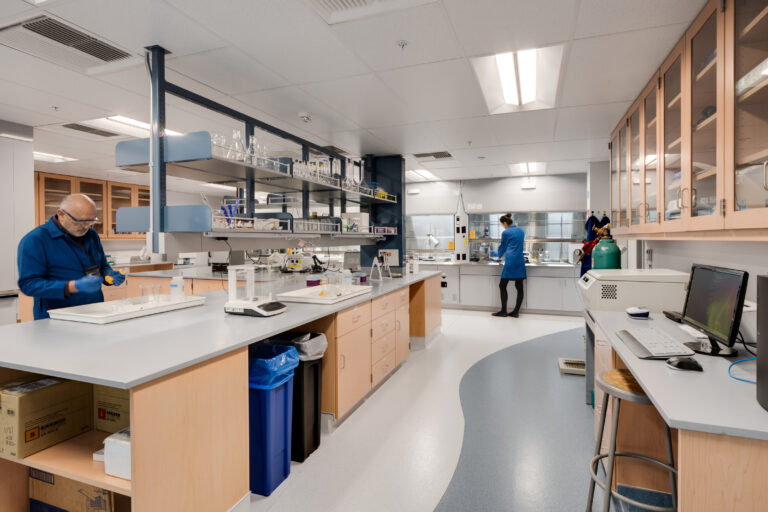 Two scientists in a modern laboratory, working at benches surrounded by glassware, cabinets, and computer equipment