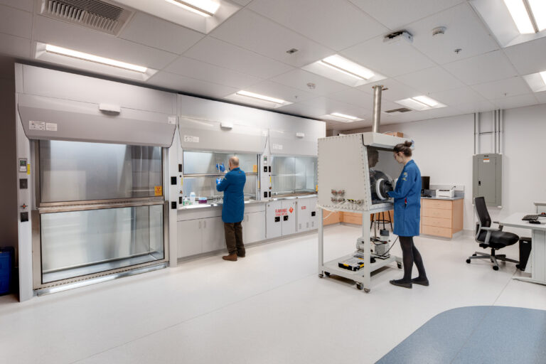 Two scientists in blue lab coats work in a clean laboratory with fume hoods and equipment
