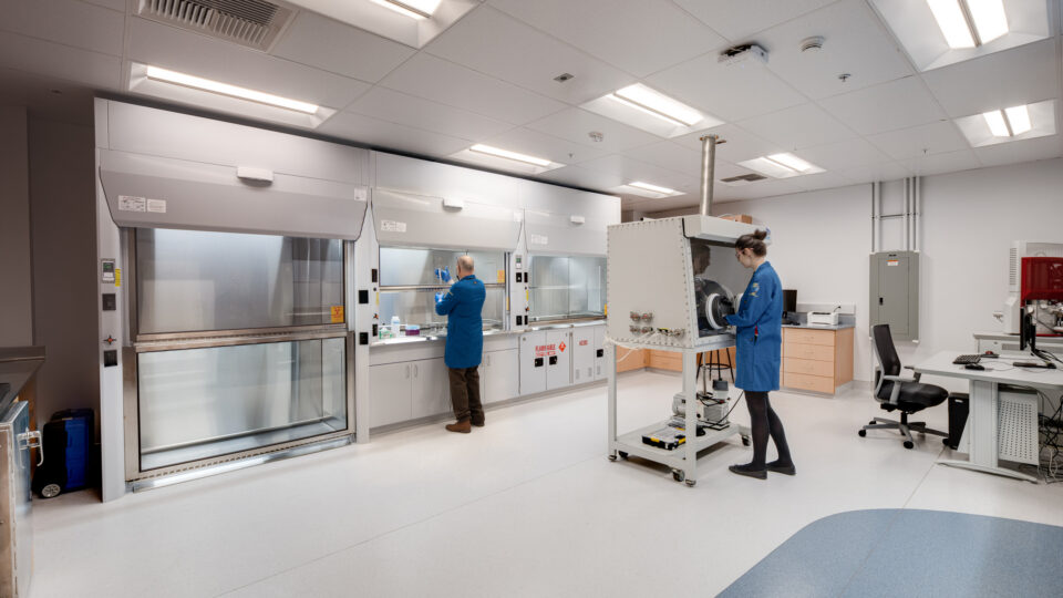 Two scientists in blue lab coats work in a clean laboratory with fume hoods and equipment