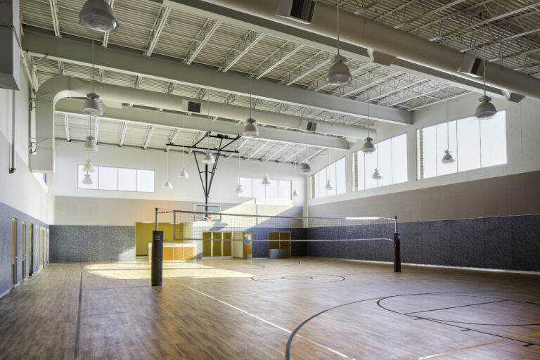 Empty indoor gym with a volleyball net, basketball hoop, and sunlit wood floor under high ceilings
