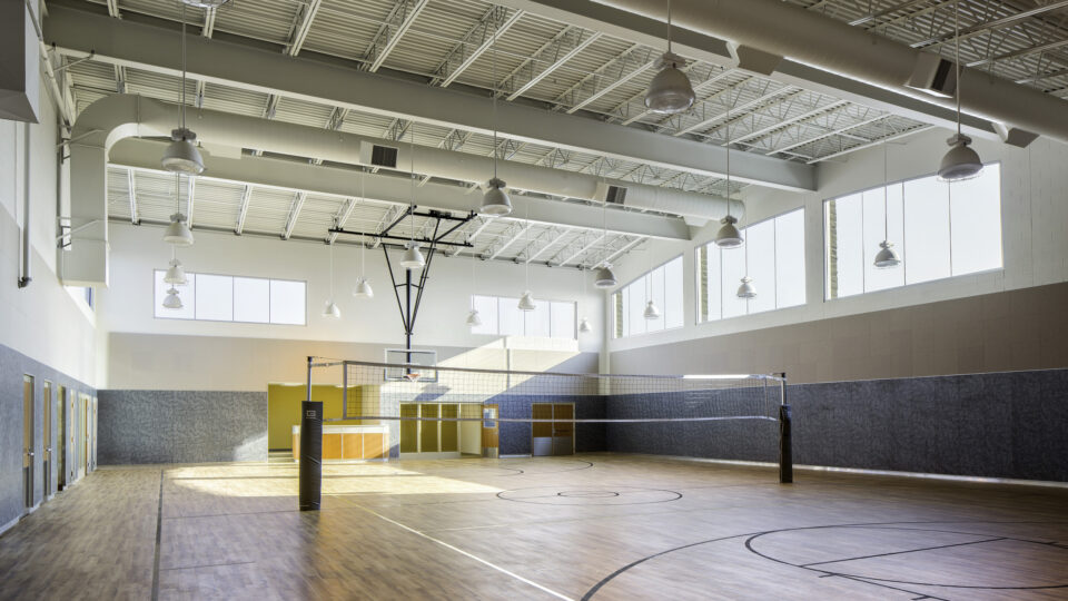 Empty indoor gym with a volleyball net, basketball hoop, and sunlit wood floor under high ceilings