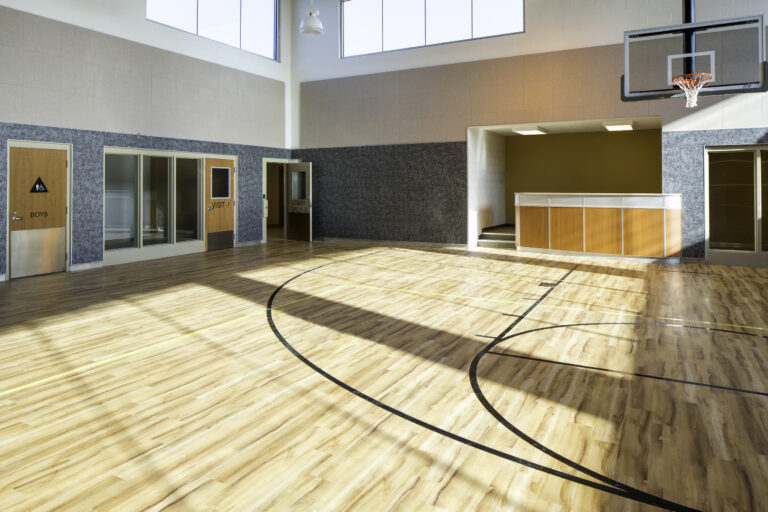 Empty indoor basketball gym with a hoop, wood floor, and doors labeled boys and visitor