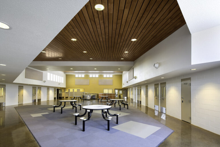 Modern school cafeteria with round tables, a wood-paneled ceiling, and classroom doors along the walls