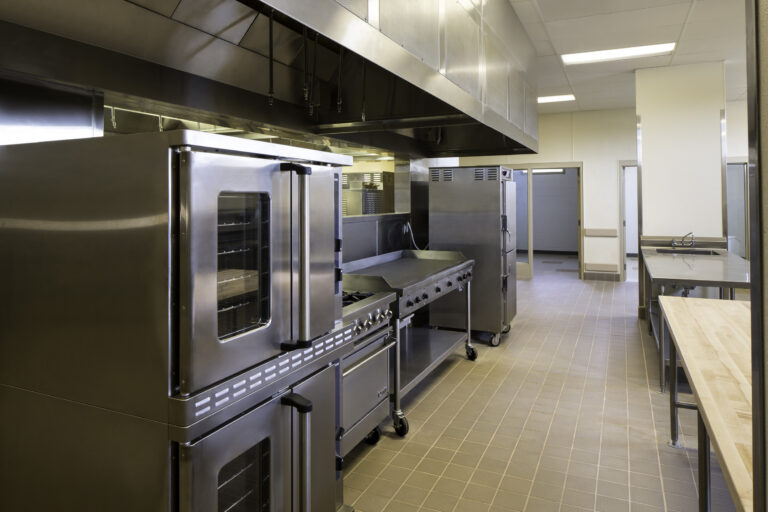 Empty stainless steel commercial kitchen with ovens, prep tables, and overhead ventilation hood