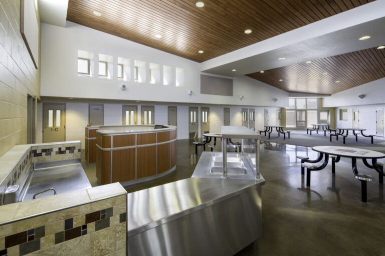 Empty institutional cafeteria with stainless-steel serving counters, round tables, and a wood-paneled ceiling with skylights